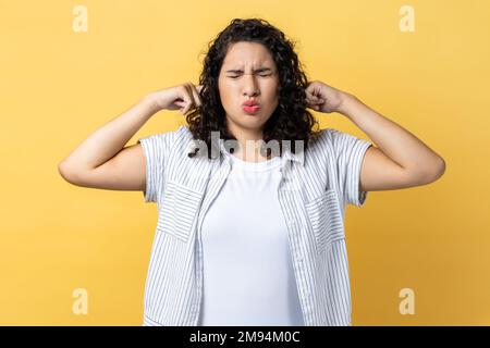 Don't want to listen. Portrait of nervous woman with dark wavy hair irritated by loud noise covering ears and grimacing in pain. Indoor studio shot isolated on yellow background. Stock Photo