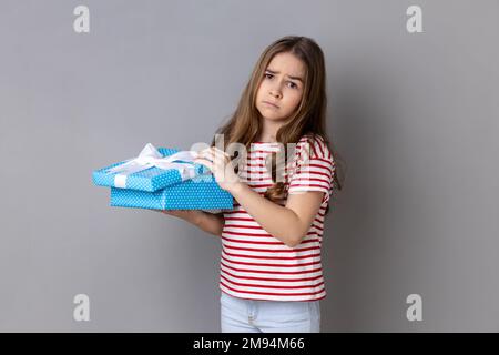 Portrait of little girl wearing striped T-shirt opening birthday gift box and looking at camera with disappointed expression, unwrapping bad present. Indoor studio shot isolated on gray background. Stock Photo