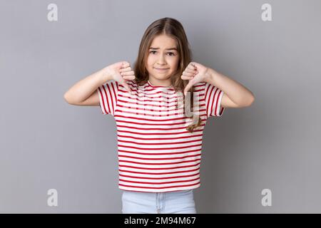 Portrait of unhappy sad little dark haired girl wearing striped T-shirt showing thumb down, expressing bad negative emotions, dislike. Indoor studio shot isolated on gray background. Stock Photo