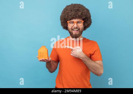 Portrait of smiling man with Afro hairstyle wearing orange T-shirt and funny eyeglasses holding pointing at paper house, helping with rent. Indoor studio shot isolated on blue background. Stock Photo