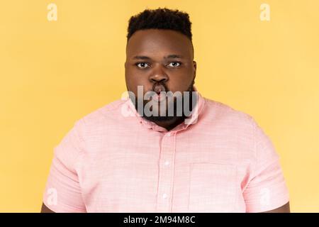 Portrait of funny positive young adult man wearing pink shirt standing with pouted lips making fish face, having fun, childish good mood. Indoor studio shot isolated on yellow background. Stock Photo