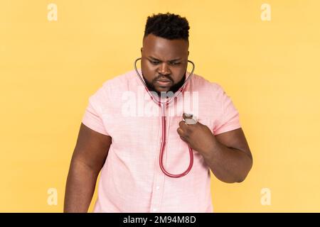 Portrait of concentrated bearded man wearing pink shirt standing with stethoscope, doing self examining of his heart, medicine concept. Indoor studio shot isolated on yellow background. Stock Photo