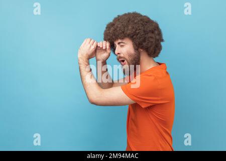 Side view of man with Afro hairstyle wearing orange T-shirt making glasses shape, looking through monocular gesture with amazed expression. Indoor studio shot isolated on blue background. Stock Photo