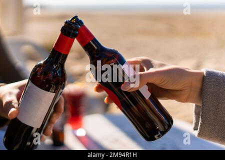 Crop anonymous male and female clinking bottles of beer while celebrating occasion on blurred background of seashore Stock Photo