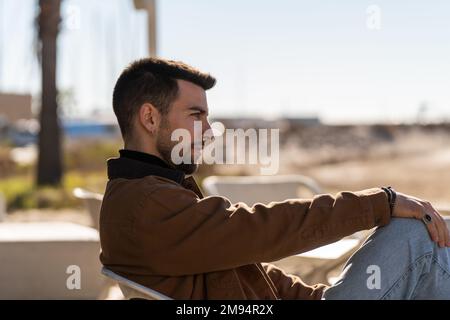 Side view of serious male in outerwear sitting on chair against blurred background of street in sunny weather Stock Photo