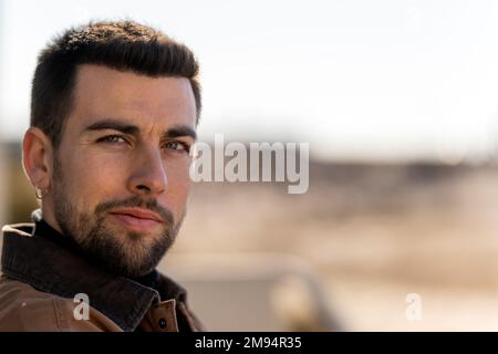 Portrait of positive bearded male with dark hair and earring in warm clothes looking at camera while sitting against blurred background Stock Photo