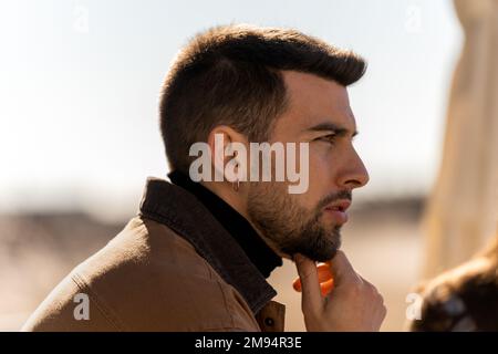 Side view of pensive bearded guy in outerwear with earring looking away thoughtfully while touching chin on blurred background Stock Photo