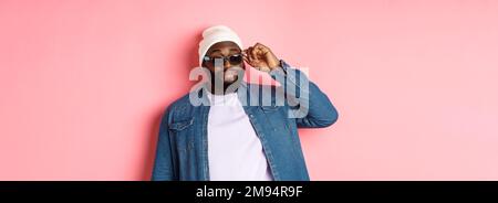 Cool and sassy african-american bearded man, looking confident, touching sunglasses and stare at camera please, standing over pink background Stock Photo