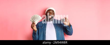 Dreamy african-american man showing credit card and dollars, thinking about shopping and smiling, standing over pink background Stock Photo