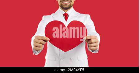 Close up of red heart shaped Valentine card in hands of happy young man in white suit Stock Photo