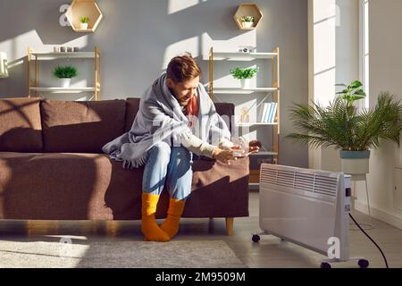 Man in socks and blanket warms his hands by a heater in a cold room at home in winter Stock Photo