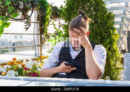 Upset man looking at smartphone holding his head while sitting in penthouse got bad news Stock Photo