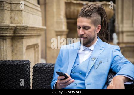 Young successful man in a blue suit looking at a smartphone while sitting in a luxury street cafe Stock Photo