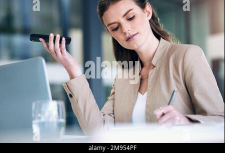 Playing back a voice message from her colleague. a young businesswoman writing notes while using a cellphone in an office. Stock Photo