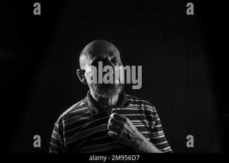 A grayscale of a handsome bald mature man with a beard wearing a casual shirt on a black background Stock Photo