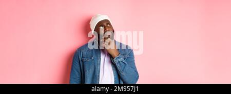 Image of puzzled and concerned black man, staring at upper left corner and thinking, making decision with doubts, standing over pink background Stock Photo