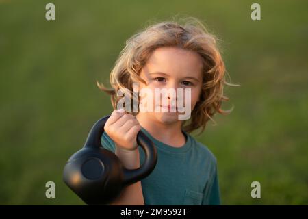 Child lifting the kettlebell in park outside. Child boy pumping up biceps muscles with kettlebell. Fitness kids with dumbbells Stock Photo