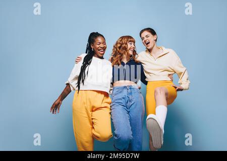 Group of cheerful female friends having fun while embracing each other. Three happy young women laughing and having a good time while standing against Stock Photo