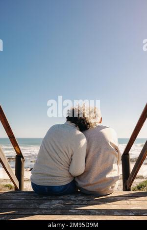 Back view of an elderly couple getting a refreshing view of the ocean while sitting on a wooden foot bridge. Retired senior couple spending some quali Stock Photo