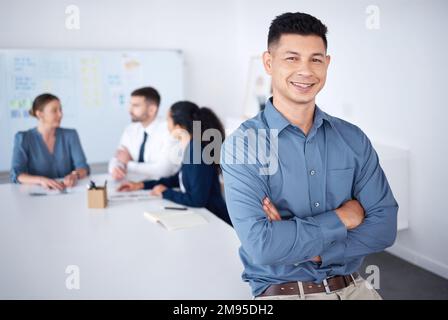 Portrait of confident smiling mixed race businessman leaning on desk in a boardroom with arms folded. Diverse group of businesspeople in meeting and Stock Photo