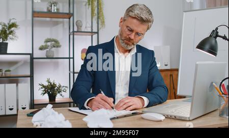Angry furious mature man working at office throwing crumpled paper, having nervous breakdown at work, migaine, headache, stress management, mental distress problems, losing temper, reaction on failure Stock Photo
