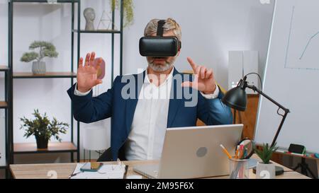 Mature businessman programmer software developer using headset helmet app to play simulation game. Watching virtual reality 3D 360 video. Mature old gray-haired in VR goggles working at home office Stock Photo