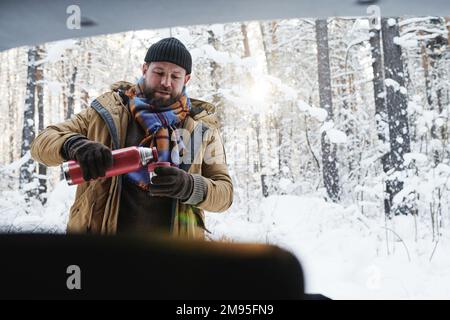 Young bearded man in warm clothing drinking hot tea during picnic in winter forest Stock Photo