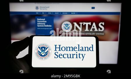 Person holding mobile phone with seal of American Department of Homeland Security (DHS) on screen in front of web page. Focus on phone display. Stock Photo