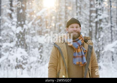 Portrait of young bearded man in warm clothing smiling at camera standing in winter forest Stock Photo