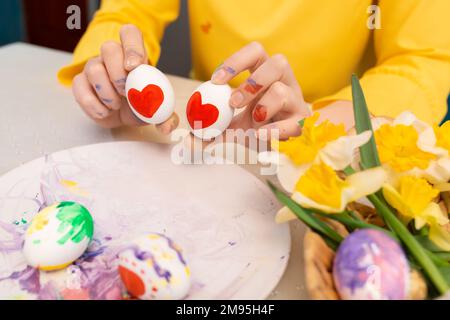 Female hands holding two painted with hearts shapes eggs near plate with easter decorations. Concept of creativity and love. Stock Photo