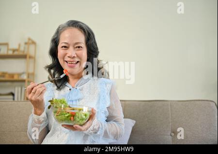 Healthy and cheerful Asian 60s aged woman enjoys eating tasty Buddha salad vegetables mix bowl in her living room. healthy lifestyle concept Stock Photo