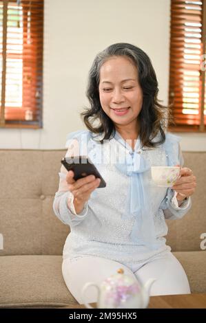 Portrait, Charming and happy Asian retired woman using her mobile phone on sofa while enjoying afternoon tea brunch in her living room. Stock Photo