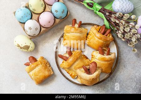 Buns in the form of an Easter rabbit from sausage and cheese in a yeast dough with colored eggs on a gray stone background. Traditional Easter symbols Stock Photo