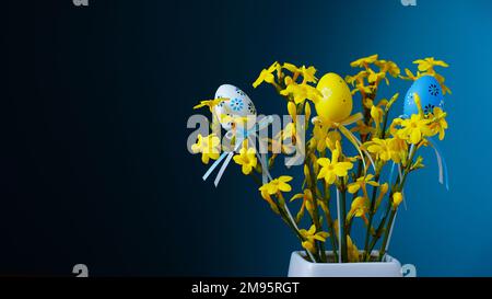 Happy Easter. A bouquet of spring yellow flowers on blue background with easter eggs with pattern in a white vase. Stock Photo