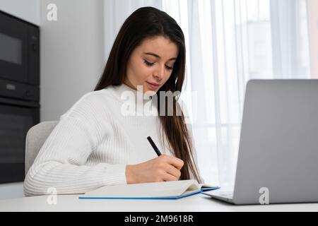 Intelligent female student writing notes to textbook sitting at home and preparing to the exam.  Stock Photo