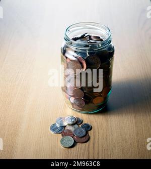 An old glass jar filled with savings of lots of coins on a wooden table top. Some of the coins are on the table Stock Photo