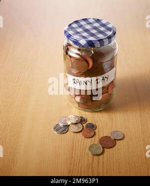 An old glass jar filled with savings of lots of coins on a wooden table top. Some of the coins are on the table Stock Photo