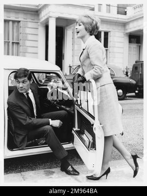 A man sits in the driver's seat of a car while a woman leans on the open door. Stock Photo