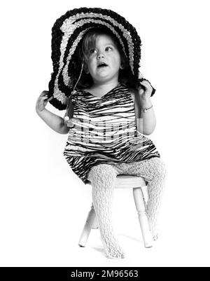A little girl sitting on a stool wearing a fancy hat. Stock Photo
