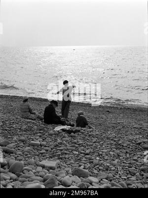 Skimming stones. A young boy, standing next to his seated family, flings flat stones into the sea from the pebble beach, trying to make them skim across the surface in the evening light. Photograph by Norman Synge Waller Budd Stock Photo