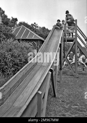 Two children, a girl and a boy, with two women at the top of a wooden playground slide in Bexhill, Sussex.  Horses on a roundabout can be seen in the background. Stock Photo