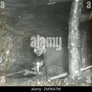 A miner undercutting or holing the coal in a narrow seam, Plas y Coed Level, South Wales.  A solitary candle provides him with light. Stock Photo