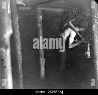 A miner, stripped to the waist, with a pick resting across his shoulder and a lamp in his hand, in a coal seam in South Wales. Stock Photo