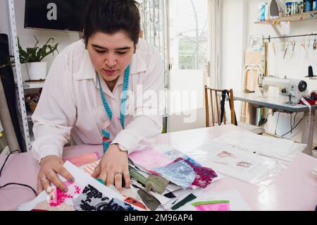 Caucasian young woman working at home in her clothing design workshop, looking for fabric samples in the catalog on the table, textile industry concep Stock Photo