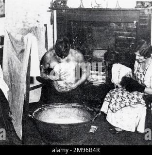 A South Wales miner washes at a tin bath in front of the fire, while his wife sits mending socks. Stock Photo