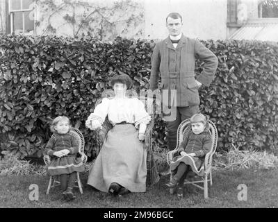 A middle class Edwardian family (parents and two daughters) pose for a photograph in their garden, probably in the Mid Wales area.  Mother and daughters are sitting on wickerwork chairs.  The little girls are wearing identical dresses. Stock Photo