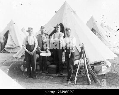 A group of four soldiers at a training camp, informally dressed, posing for their photo outside a tent.   A stook of rifles can be seen on the right. Stock Photo
