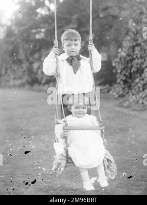 Two Edwardian children on a swing in a garden, Mid Wales.  The swing has a safety bar at the front. Stock Photo