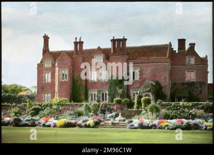 An unidentified garden in Cambridgeshire with a view across the lawn, showing colourful plants and shrubs growing on either side of a flight of stone steps, a red brick garden wall, and a large red brick house with tall chimneys in the background. Stock Photo