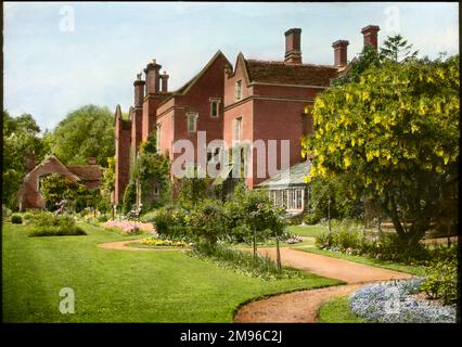 An unidentified garden in Cambridgeshire, showing a large red brick house with tall chimneys, a greenhouse, a well-kept lawn, trees, flowers and shrubs, and a garden path. Stock Photo